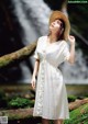 A woman in a white dress and straw hat standing in front of a waterfall.